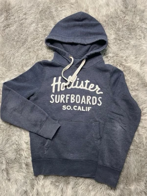 Blå Hollister hoodie med tryck - Mysig blå hoodie från Hollister med vit text 'Hollister Surfboards SO. CALIF' på bröstet. Klassisk känguruficka framtill, ribbade muddar och justerbar huva med vita snören. Perfekt för en avslappnad och cool stil.