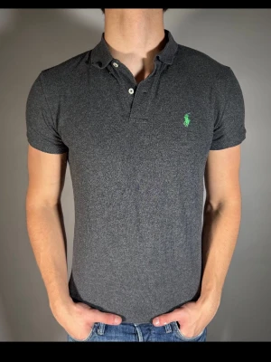 Mörkgrå pikétröja från Ralph Lauren - Snygg mörkgrå pikétröja från Ralph Lauren med klassisk krage och två knappar framtill. Tröjan har en grön broderad logga på bröstet och är tillverkad i mjuk bomull. Perfekt för en stilren och avslappnad look.