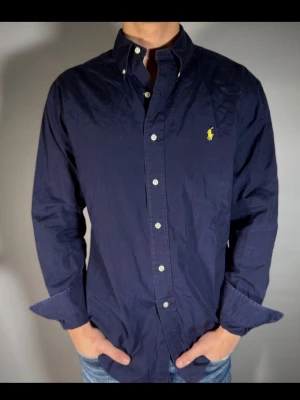 Mörkblå skjorta från Ralph Lauren - Klassisk mörkblå skjorta från Ralph Lauren med knappslå och ikonisk gul broderad logga på bröstet. Skjortan har lång ärm, normal passform och är tillverkad i bomull. Perfekt för en stilren och avslappnad look.