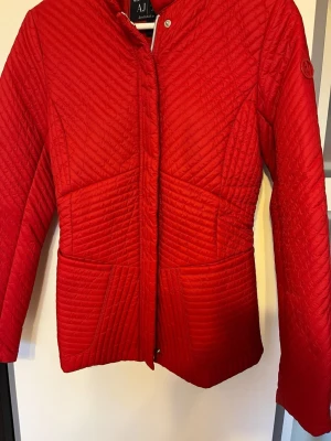 Röd tunn jacka från Armani Jeans - Snygg röd tunn jacka från Armani Jeans med coolt quiltat mönster och figurnära passform. Jackan har dragkedja framtill, två stora fickor och diskret logga på ärmen. Perfekt för kalla vinterdagar och ger en riktigt fräsch look.