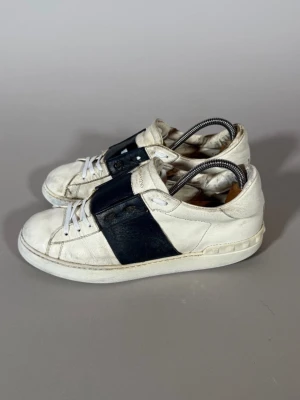 Valentino vita sneakers med svart detalj - Snygga vita sneakers från Valentino med en bred svart läderdetalj över sidan och tungan. Klassisk låg modell med vita snören och rundad tå. Skorna har en stilren och modern look som passar till det mesta. Tillverkade i läder för en exklusiv känsla.