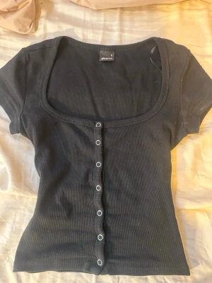 Söt Gina tricot topp - Säljer en svart ribbad topp från Ginatricot med korta ärmar och djup, rundad urringning. Toppen har knappar framtill och sitter tight. Perfekt till jeans eller kjol för en enkel men snygg look.