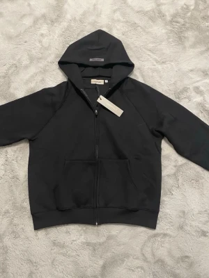 Svart Essentials hoodie med dragkedja - Säljer en svart hoodie från Essentials med dragkedja framtill och stor ficka på magen. Hoodien har en klassisk huva och Essentials-tryck på ryggen. Tillverkad i mjukt bomullsmaterial som är perfekt för en avslappnad streetwear-look.