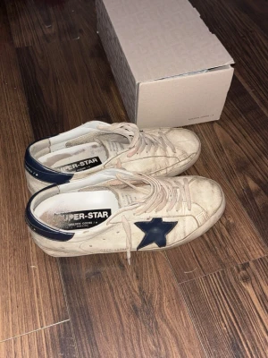 Golden Goose Superstar sneakers beige/blå - Säljer ett par Golden Goose Superstar sneakers i beige med mörkblå stjärna på sidan, ny pris är runt 5000 och då dom är ganska använda så säljer jag för ett rimligt pris säljer för 1149! Storlek 43. Hör av dig vid intresse! Kan eventuellt gå ner lite i pris vid en snabb affär 