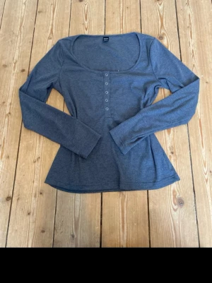 Mörkgrå långärmad topp med knappar - Mörkgrå långärmad topp med rundad, djup urringning och dekorativa knappar framtill. Figurnära passform och mjukt material som känns skönt mot huden. Perfekt att styla med jeans eller kjol för en enkel men snygg look.