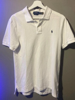 Vit pikétröja från Polo Ralph Lauren - Klassisk vit pikétröja från Polo Ralph Lauren med korta ärmar och blå broderad logga på bröstet. Tröjan har krage och knappar vid halsen. Perfekt till jeans eller shorts för en clean och stilren look.