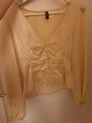 Champagnefärgad/gul blus från H&M - Snygg och populär blus  med v-ringning och knappar framtill. Blusen har en liten fläck på ena ärmen, men det är inget som syns tydligt. Materialet är i satin och blusen passar perfekt till våren och sommaren.💗