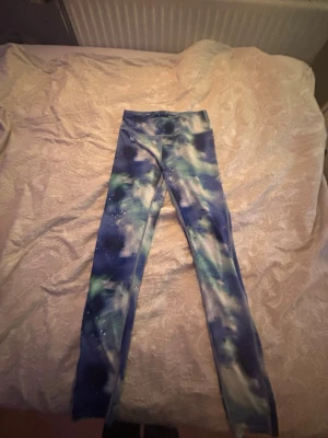 Blå leggings med galaxmönster - Säljer ett par stretchiga leggings med blått och vitt galaxmönster. De har hög midja och är tillverkade i ett mjukt, följsamt material som passar perfekt till både träning och chill. Mönstret har inslag av grönt och svart för en unik look.