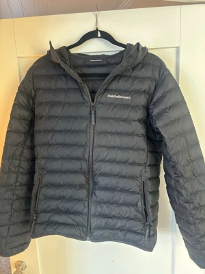 Svart dunjacka Peak Performance M - Snygg svart dunjacka från Peak Performance med huva och dragkedja framtill. Jackan har två sidofickor med dragkedja och diskret logga på bröstet. Perfekt för kalla vinterdagar och riktigt lätt att bära.