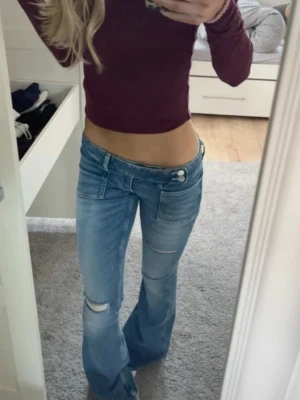 Ljusblå low waist bootcut jeans - Säljer ett par ljusblå low waist jeans med bootcut passform och slitningar vid knäet. Jeansen har dubbla knappar i linningen och klassiska fem fickor. Perfekta för en trendig och avslappnad look.