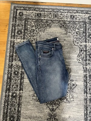 Blå skinny jeans från Tommy Hilfiger - Säljer ett par blå skinny jeans från Tommy Hilfiger med klassisk femficksdesign och logga på bakfickan. Jeansen har en snygg tvättad look och smal passform som sitter tajt längs benen. Perfekta för en trendig och avslappnad stil.