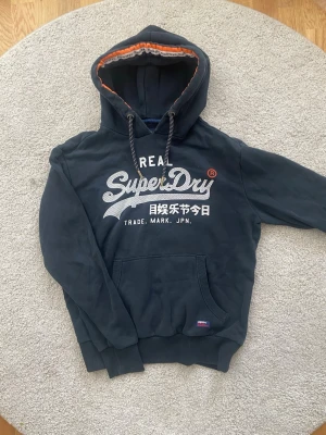 Mörkblå Superdry hoodie med tryck - Snygg mörkblå hoodie från Superdry med stort vitt tryck och japanska tecken på bröstet. Hoodien har en klassisk känguruficka, dragsko i huvan och orange detalj på insidan av huvan. Perfekt för en avslappnad streetwear-look.