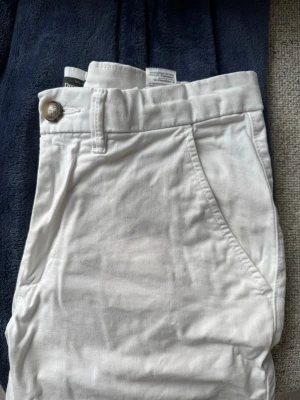 Vita shorts i bomull med fickor - Snygga vita shorts med normal passform och klassisk design. Shortsen har två sidofickor, bälteshällor och stängs med knapp och dragkedja. Tillverkade i mjuk bomull, perfekta för varma dagar.