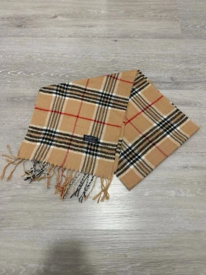 Beige Burberry Scarf – 100% Lambswool – Made in England - Klassisk beige halsduk från Burberry med ikoniskt rutmönster i svart, vitt och rött. Halsduken har fransar i ändarna och är tillverkad i mjukt material, perfekt för att ge din outfit en tidlös touch.