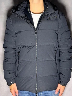 J.Linderberg jacka - Snygg mörkgrå pufferjacka med dragkedja framtill och två sidofickor med dragkedja. Jackan har en hög krage, avtagbar huva och quiltad design. Perfekt för kalla vinterdagar och har en modern, stilren look. Bara höra av dig vid frågor eller liknande