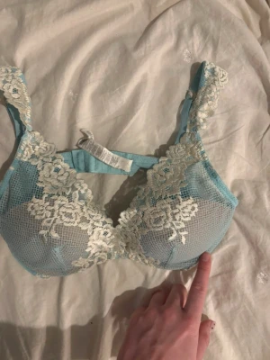 Intimissimi BH - Säljer en ljusblå intimissimi bh med vacker vit spets och blommönster. Bh:n har breda axelband dekorerade med spets och bygel för extra stöd. Materialet är genomskinligt mesh med detaljer i spets.