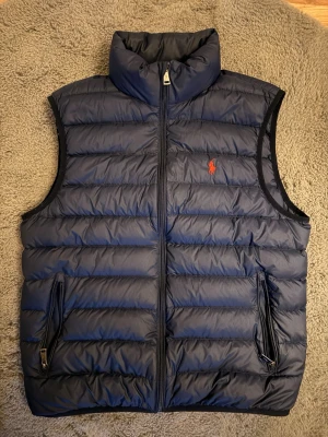 Polo Ralph Lauren Väst - Snygg mörkblå dunväst från Polo Ralph Lauren med klassisk quiltad design och hög krage. Västen har två dragkedjeförsedda fickor och den ikoniska röda loggan broderad på bröstet. Perfekt för lager på lager och riktigt stilren look.