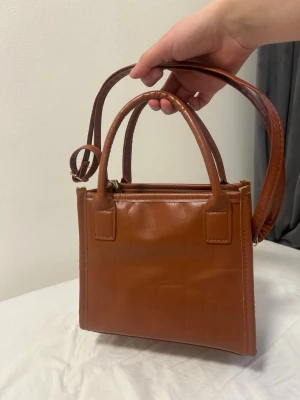 Brun fyrkantig handväska i fejkläder - Snygg brun handväska i fyrkantig modell med både handtag och justerbar axelrem. Väskan är gjord i fejkläder med croco-mönster och har ett rymligt huvudfack. Perfekt för dig som gillar stilrena accessoarer med trendig touch.