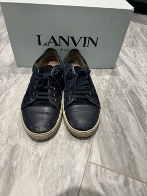 Marinblå sneakers från Lanvin - Snygga marinblå sneakers från Lanvin med ovandel i mocka och tåparti i slätt läder. Skorna har vita gummisulor och klassisk snörning. Perfekta för dig som gillar stilrena och exklusiva sneakers med en lyxig känsla.