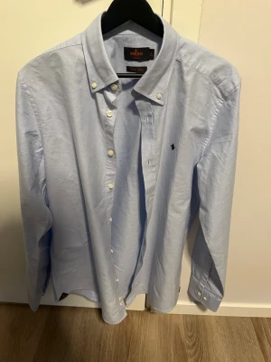Ljusblå skjorta från Morris - Snygg ljusblå skjorta från Morris i slim fit-modell. Skjortan har klassisk button-down krage, vita knappar och broderad logga på bröstet. Tillverkad i bomull och passar perfekt till en stilren look.