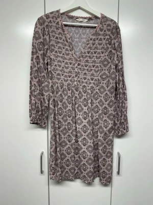 Mönstrad klänning Odd Molly - Supersöt kortklänning från Odd Molly med blommigt mönster i ljusrosa och grå toner. Klänningen har v-ringning, knappar framtill och trekvartsärm. Tillverkad i mjuk bomull och dekorerad med virkade detaljer vid halsen. Perfekt för dig som gillar boho-stil!