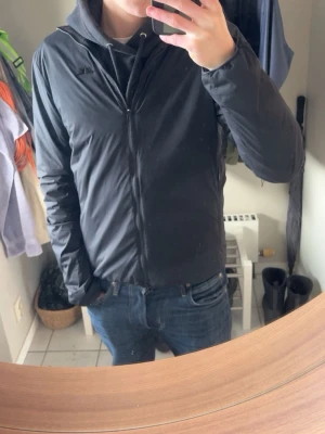 Svart vindjacka från J.Lindeberg - Snygg svart vindjacka från J.Lindeberg med minimalistisk design och diskret logga på bröstet och ryggen. Jackan har dragkedja framtill, huva och smal passform. Perfekt för blåsiga dagar och enkel att matcha med olika outfits.    Passar även bra till golfen.                                               Model: Clinton                                                                       Priset är inte hugget i sten:)