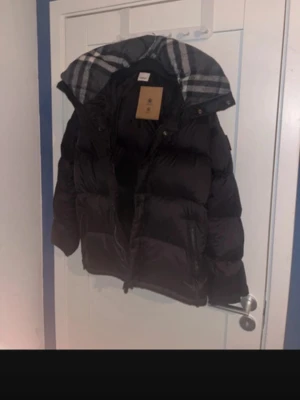 Burberry jacka/väst - Snygg svart pufferjacka med stor vaddering och huva. Jackan har dragkedja framtill och rutigt foder i huvan för extra stil. Perfekt för kalla vinterdagar och ger en riktigt trendig look. Passar dig som vill ha både värme och streetstyle.