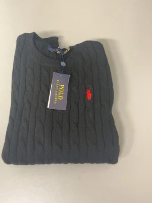 Svart kabelstickad tröja Polo Ralph Lauren - Kabelstickad svart tröja från Polo Ralph Lauren med rund halsringning och röd broderad logga på bröstet. Tröjan har ribbade muddar vid ärmslut och nederkant. Klassisk och stilren design som passar till mycket.