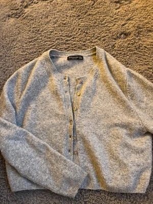 Grå kofta från Stradivarius - Mysig grå kofta från Stradivarius med rund halsringning och knappar framtill. Koftan har långa ärmar och ribbade muddar vid ärmslut och nederkant. Perfekt att slänga över en t-shirt eller linne för en chill look.