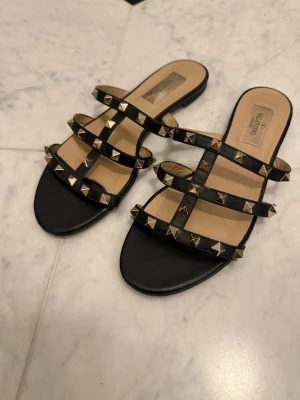 Svarta sandaler med nitar från Valentino - Säljer ett par svarta sandaler från Valentino med flera remmar dekorerade med guldfärgade nitar. Sandalerna har öppen tå och platt sula, tillverkade i läder med beige innersula och cool rockig vibe.Använda flitigt kan lätt sulas om/ÄKTA! 