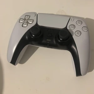 Sony DualSense PS5 handkontroll vit helt ny - Säljer en vit och svart Sony DualSense handkontroll till PlayStation 5. Kontrollern har en futuristisk design med touchpad, två analoga spakar, klassiska PlayStation-knappar och triggers. Tillverkad i plast med matt finish och ergonomiskt grepp.