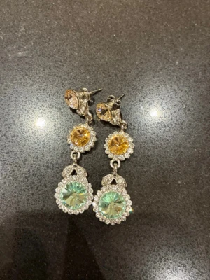 Glittriga hängande örhängen med stenar - Snygga örhängen med tre stora gnistrande stenar i gult och ljusgrönt, omgivna av små klara stenar. Örhängena är silvriga och har en droppformad design som ger en lyxig känsla. Perfekta för dig som vill sticka ut med lite extra bling.