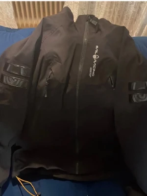 Svart vindjacka Sail Racing - Svart vindjacka från Sail Racing med dragkedja framtill och logga på bröstet samt ryggen. Jackan har två fickor med dragkedja och detaljer i svart på ärmarna. Materialet är troligtvis syntet, perfekt för blåsiga dagar. Snygg och sportig design.