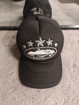 Svart Corteiz keps - Säljer en svart truckerkeps med vit brodyr framtill som föreställer ett skepp och fem stjärnor. Kepsen har mesh på sidorna och justerbar snapback. Perfekt för dig som vill ha en unik och stilren accessoar.