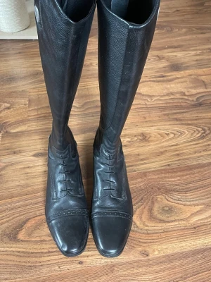 Svarta ridstövlar från CRW i läder - Snygga svarta ridstövlar från CRW i äkta läder med klassisk snörning framtill och dekorativ söm över tån. Hög modell med spänne och metallogo på skaftet. Perfekta för dig som gillar stilrena och tidlösa boots.