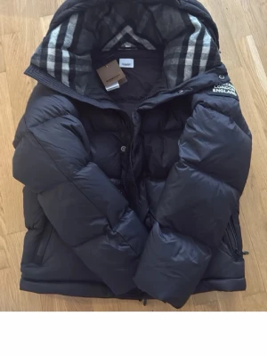 Svart pufferjacka från Burberry - Säljer en svart pufferjacka från Burberry med ikoniskt rutigt foder i huvan och tydlig logga på ärmen. Jackan har dragkedja, stora fickor och en riktigt puffig look. Perfekt för kalla dagar när du vill vara både varm och stilren.