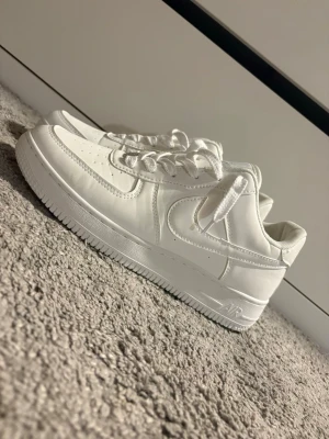 Nike Air Force 1 vita sneakers - Klassiska helvita Nike Air Force 1 sneakers i lågt snitt. Skorna har ovandel i läder, perforerad tå för ventilation och tydlig Nike Swoosh på sidorna. Tjock sula med Air-dämpning och vita skosnören. Perfekta för en clean och stilren look.