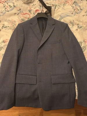 Grå kavaj med diskret rutmönster - Snygg grå kavaj med diskret rutmönster och klassisk passform. Kavajen har två fickor med lock framtill, bröstficka och helfodrad insida i mörkgrått tyg. Perfekt för en stilren och modern look.