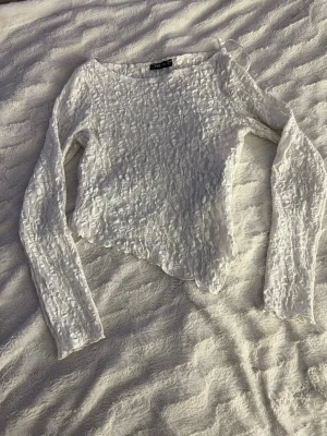 Vit spetsig långärmad topp FB Sister - Säljer en vit långärmad topp från FB Sister med spetsdetaljer och lätt skrynklig struktur. Toppen har asymmetrisk nederkant och är genombruten i materialet, vilket ger en trendig och luftig känsla.