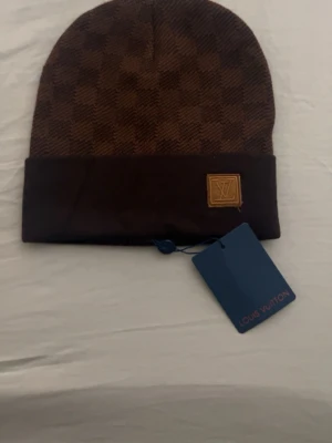 .Elegant Brun Designer Mössa – Ny med Tags - Säljer en elegant brun mössa med klassisk och stilren design. Mössan har en fin logodetalj framtill och en bekväm passform som sitter bra på huvudet.  Den bruna färgen ger ett exklusivt och tidlöst utseende som passar perfekt till många olika outfits under höst och vinter. Mössan är helt ny och säljs med tags kvar.  Detaljer: • Färg: Brun • Stilren logodetalj framtill • Ny med tags • Mjuk och bekväm passform  Perfekt för dig som vill ha en snygg och trendig mössa till kyligare dagar.