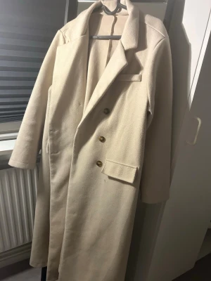 Beige lång ullrock med knappar - Stilren beige lång rock med klassisk krage och tre knappar framtill. Rocken har två fickor med lock och en bröstficka. Tillverkad i ett ull-liknande material som ger en lyxig känsla och passar perfekt till höst och vinter.