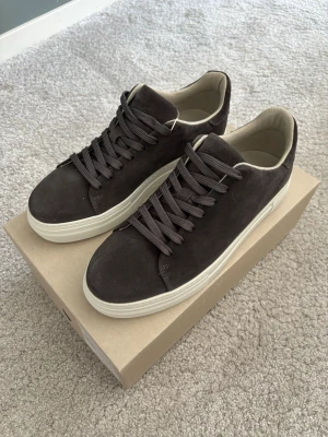 Mörkgråa Selected Homme sneakers - Stilrena mörkgrå sneakers från Selected Homme i mocka med vita sulor och matchande mörkbruna skosnören. Låg modell med beige insida och diskret design som passar till det mesta. Perfekta för dig som vill ha ett par snygga skor som går att använda till det mesta. 