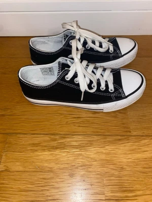 Svarta låga sneakers från 157 - Klassiska svarta låga sneakers från 157 med vit tå och vita snören. Skorna har vit sula och kontrasterande vita sömmar som ger en clean och tidlös look. Ovandelen är i canvasmaterial och sulan är i gummi. Perfekta för dig som gillar enkel och stilren design.
