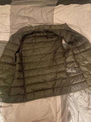 Mörkgrön dunväst från UNIQLO - Snygg mörkgrön dunväst från UNIQLO med quiltad design och hög krage. Västen har dragkedja framtill och är lätt vadderad för extra värme. Perfekt att slänga över en hoodie eller tröja för en chill och trendig look.