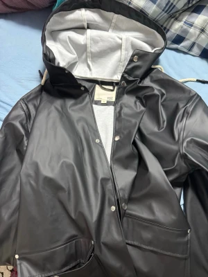 Svart regnjacka från 157 med huva - Snygg svart regnjacka från 157 med vit insida på huvan och dragsko. Jackan har tryckknappar, stora fickor framtill och är gjord i ett vattenavvisande material. Perfekt för regniga dagar och enkel att matcha med olika outfits.