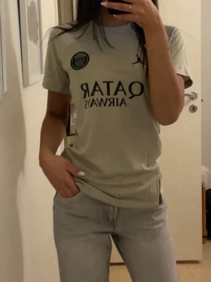 Beige PSG t-shirt med tryck - Snygg beige/grön t-shirt från PSG med svart tryck framtill, inklusive klubbmärke och texten 'Qatar Airways'. Klassisk passform och rund hals. 