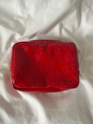 Röd necessär från YSL Beauty - Säljer en glansig röd necessär från YSL Beauty med texten 'YSL LOVE' och 'YSL BEAUTY' i små nitar på framsidan. Väskan har en rektangulär form och dragkedja upptill. Perfekt för smink eller småprylar.