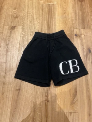 Cb shorts - Svarta shorts med bred passform och snörning i midjan. Stora vita bokstäver 'CB' broderade på ena benet ger en cool kontrast. Materialet ser ut att vara mjukt och stickat, perfekt för en avslappnad stil.