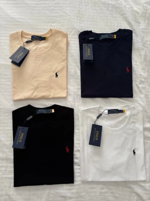 Polo Ralph Lauren t-shirt 4-pack - Fyra klassiska t-shirts från Polo Ralph Lauren i färgerna beige, marinblå, svart och vit. Alla har rund hals, diskret logga broderad på bröstet och är tillverkade i mjuk bomull. Perfekta basplagg för en clean och stilren look.