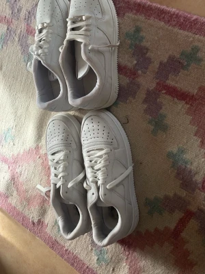 Vita Nike Air Force 1 sneakers - Klassiska helvita Nike Air Force 1 sneakers med lågt skaft. Skorna har perforerad tå, vita skosnören och den ikoniska swoosh-loggan på sidan. Tillverkade i läder med gummisula för bra grepp. Perfekta för en clean och stilren look.